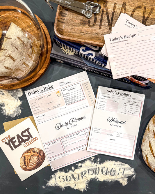 Baking-themed stationery items including a planner, notepad, and recipe cards on a dark surface with sourdough bread and flour.
