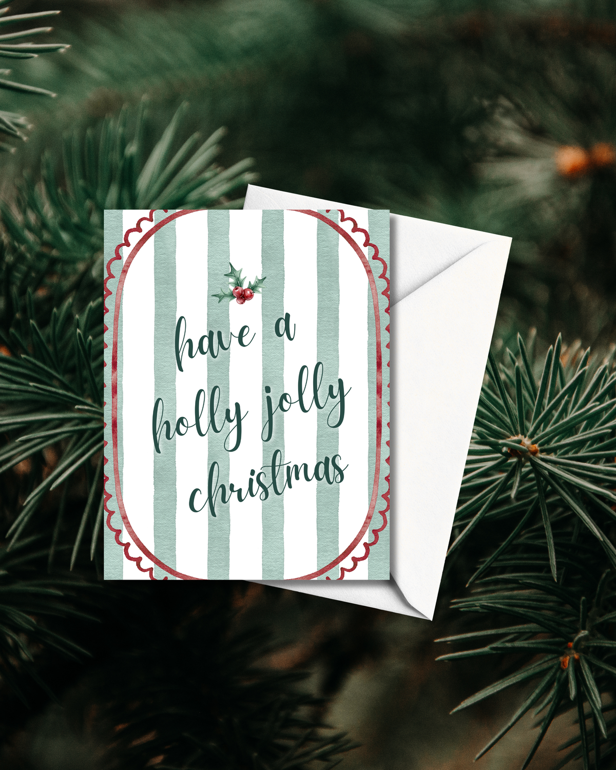 A Christmas greeting card with a white envelope, featuring the phrase 'have a holly jolly christmas' in dark teal script over pale green and white vertical stripes, with a red scalloped border and small holly sprig. The card is photographed against a blurred background of green pine needles.