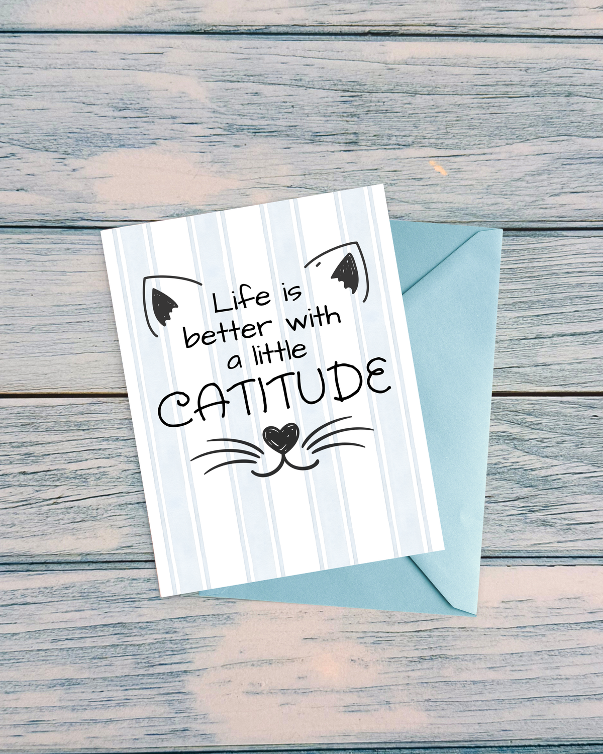 A greeting card with a light blue envelope, placed on a rustic weathered gray wooden surface. The card features a white and light blue vertical stripe background with a black cat face illustration and the text 'Life is better with a little CATITUDE'.