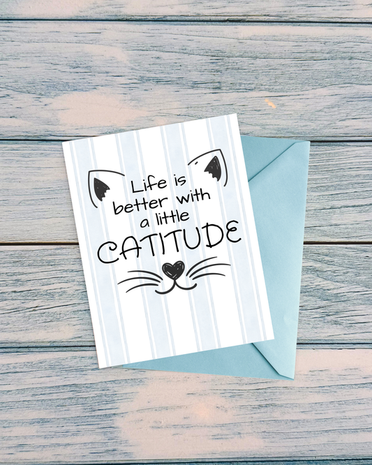 A greeting card with a light blue envelope, placed on a rustic weathered gray wooden surface. The card features a white and light blue vertical stripe background with a black cat face illustration and the text 'Life is better with a little CATITUDE'.