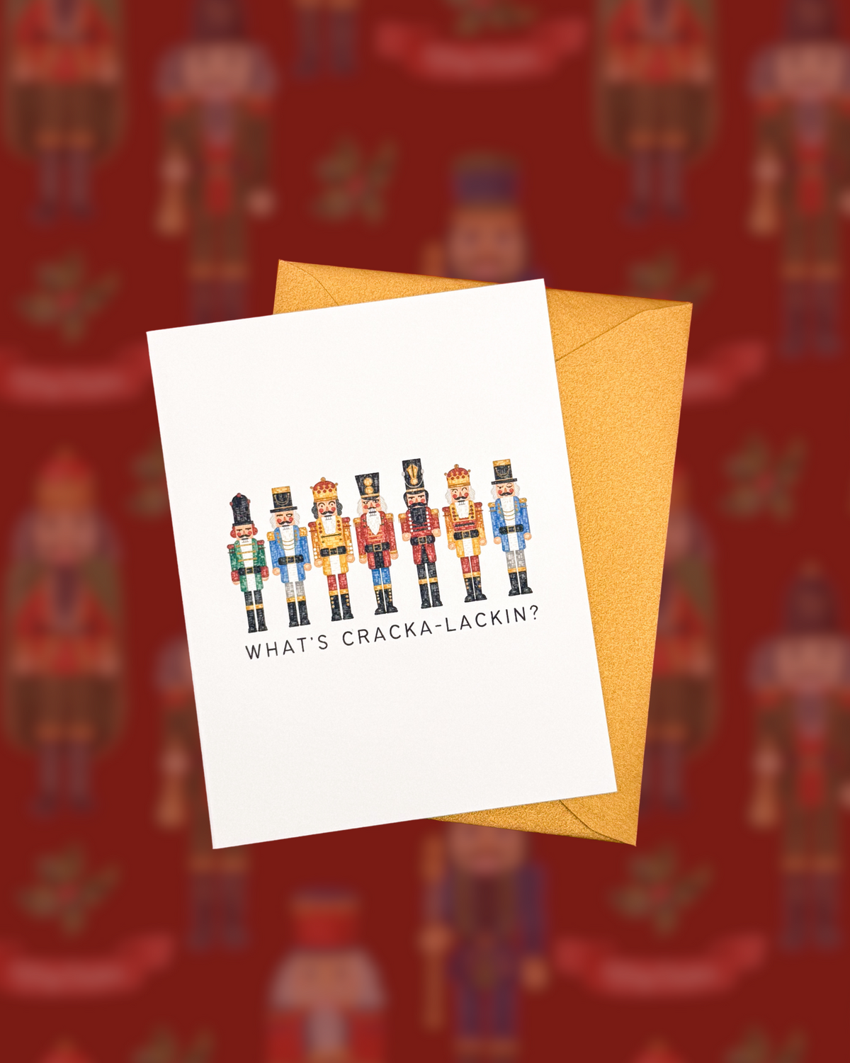 A white greeting card with a colorful illustration of nutcrackers and the text 'WHAT'S CRACKA-LACKIN?' is shown overlapping a gold envelope against a blurred red patterned background.