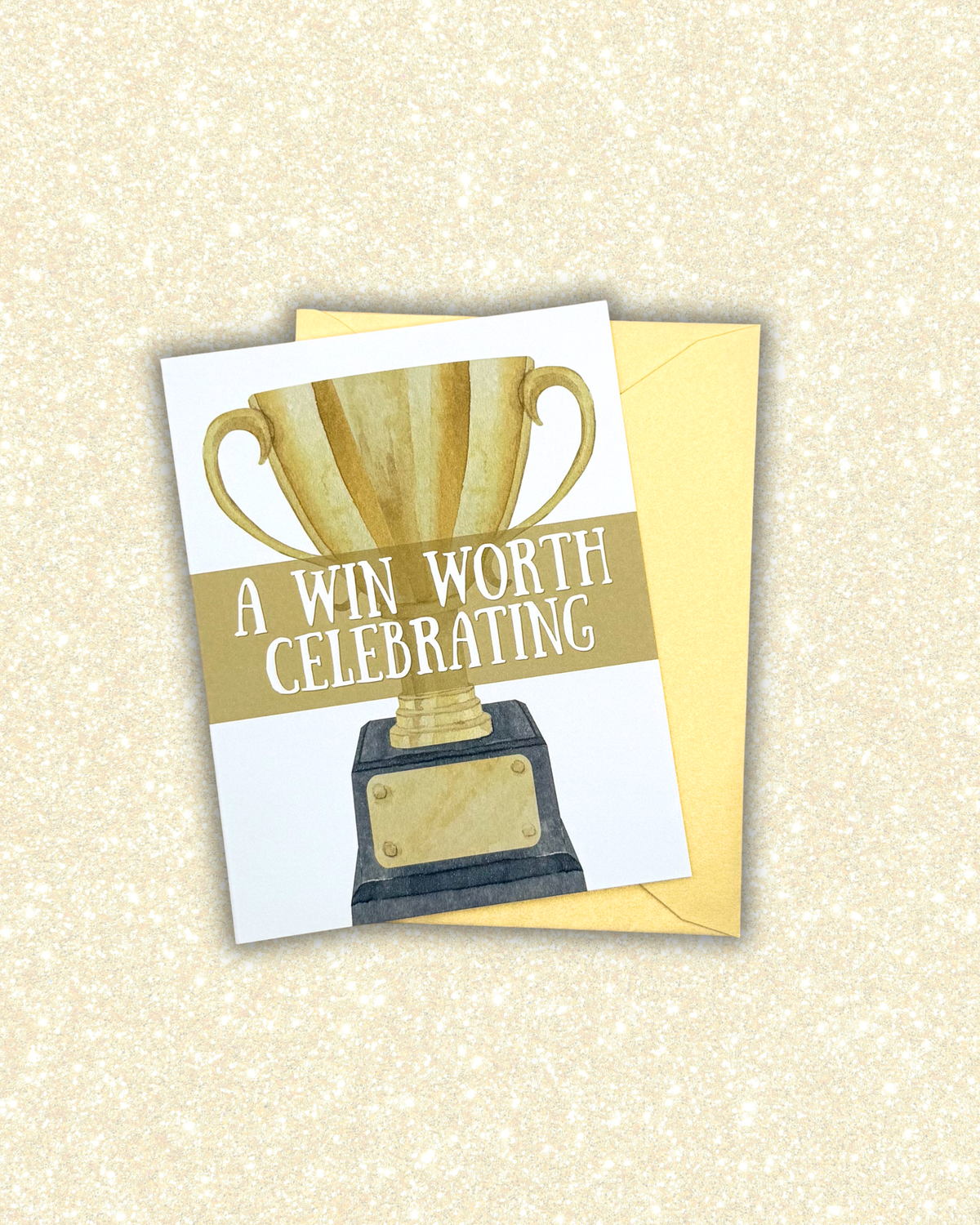 A square greeting card with a watercolor-style gold trophy and the text 'A WIN WORTH CELEBRATING,' shown with a yellow envelope on a glittery cream background.