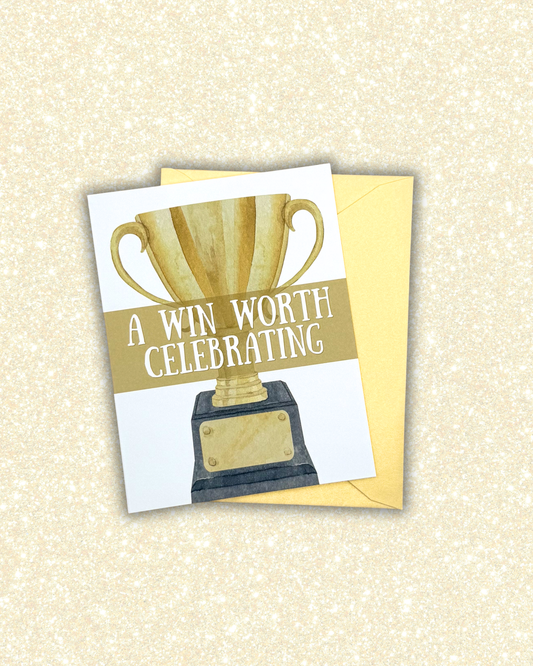 A square greeting card with a watercolor-style gold trophy and the text 'A WIN WORTH CELEBRATING,' shown with a yellow envelope on a glittery cream background.