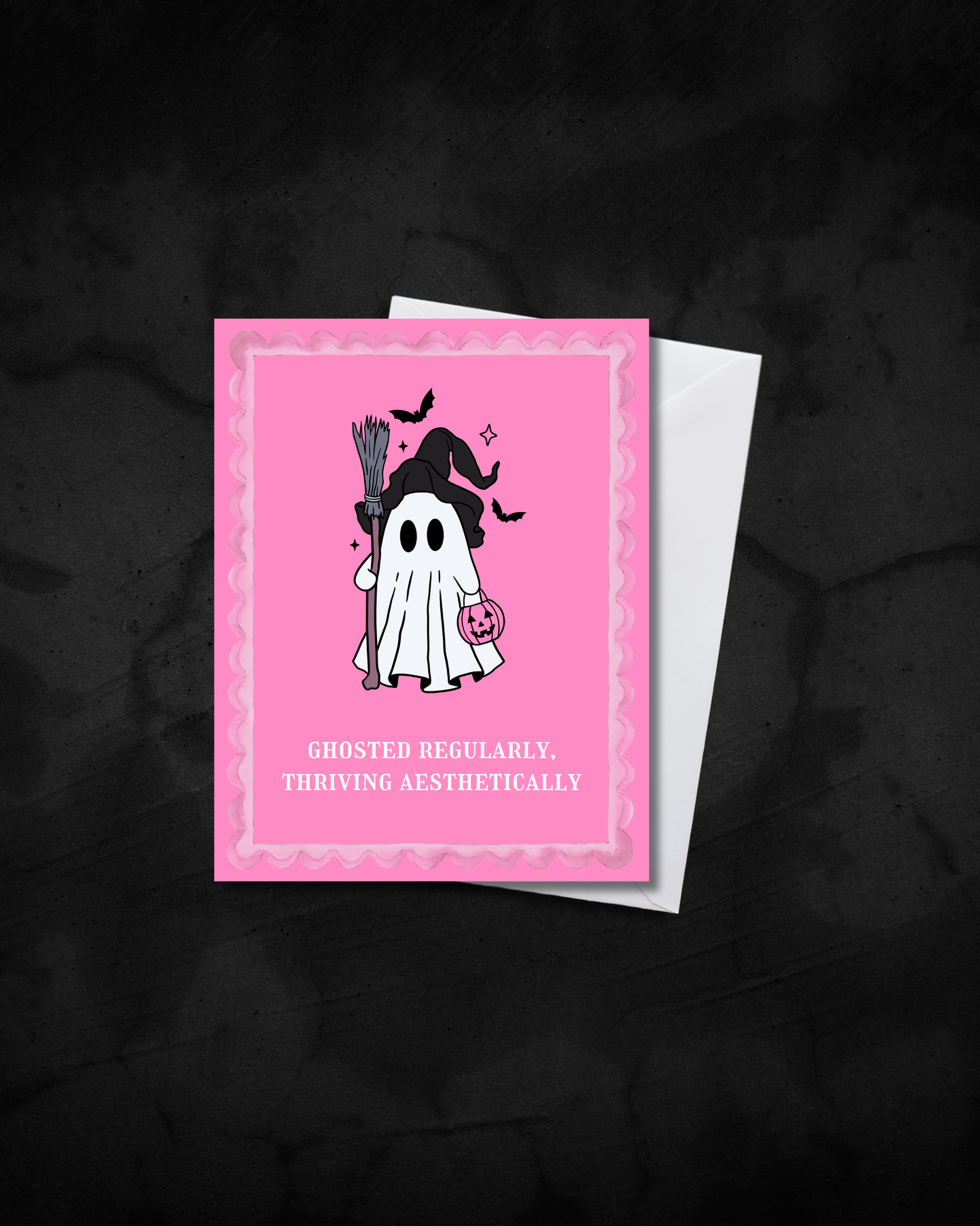 A pink Halloween greeting card with a cute ghost illustration and the text 'GHOSTED REGULARLY. THRIVING AESTHETICALLY', shown with a white envelope on a dark textured background.