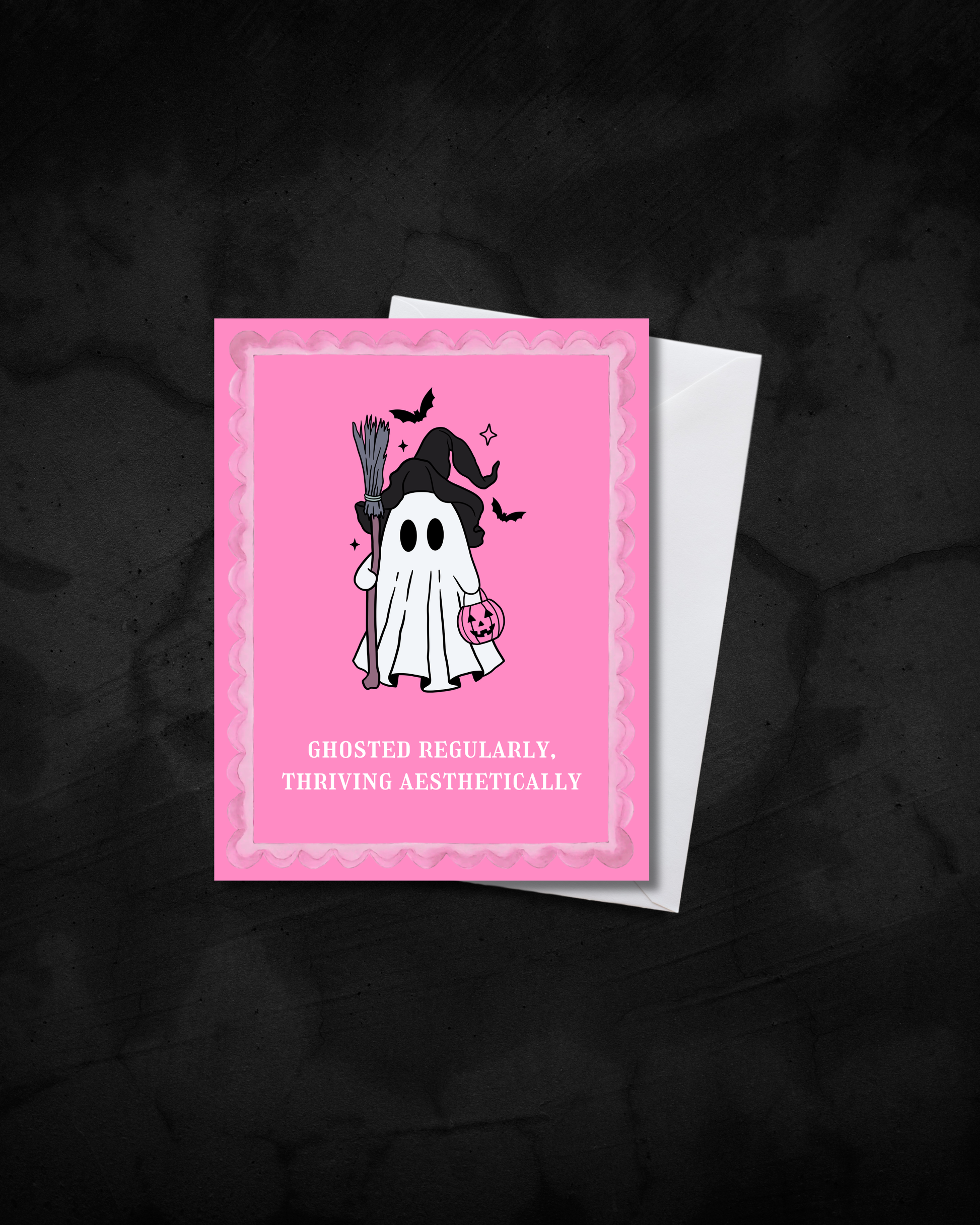 A pink Halloween greeting card with a cute ghost illustration and the text 'GHOSTED REGULARLY. THRIVING AESTHETICALLY', shown with a white envelope on a dark textured background.