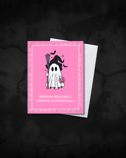 A pink Halloween greeting card with a cute ghost illustration and the text 'GHOSTED REGULARLY. THRIVING AESTHETICALLY', shown with a white envelope on a dark textured background.