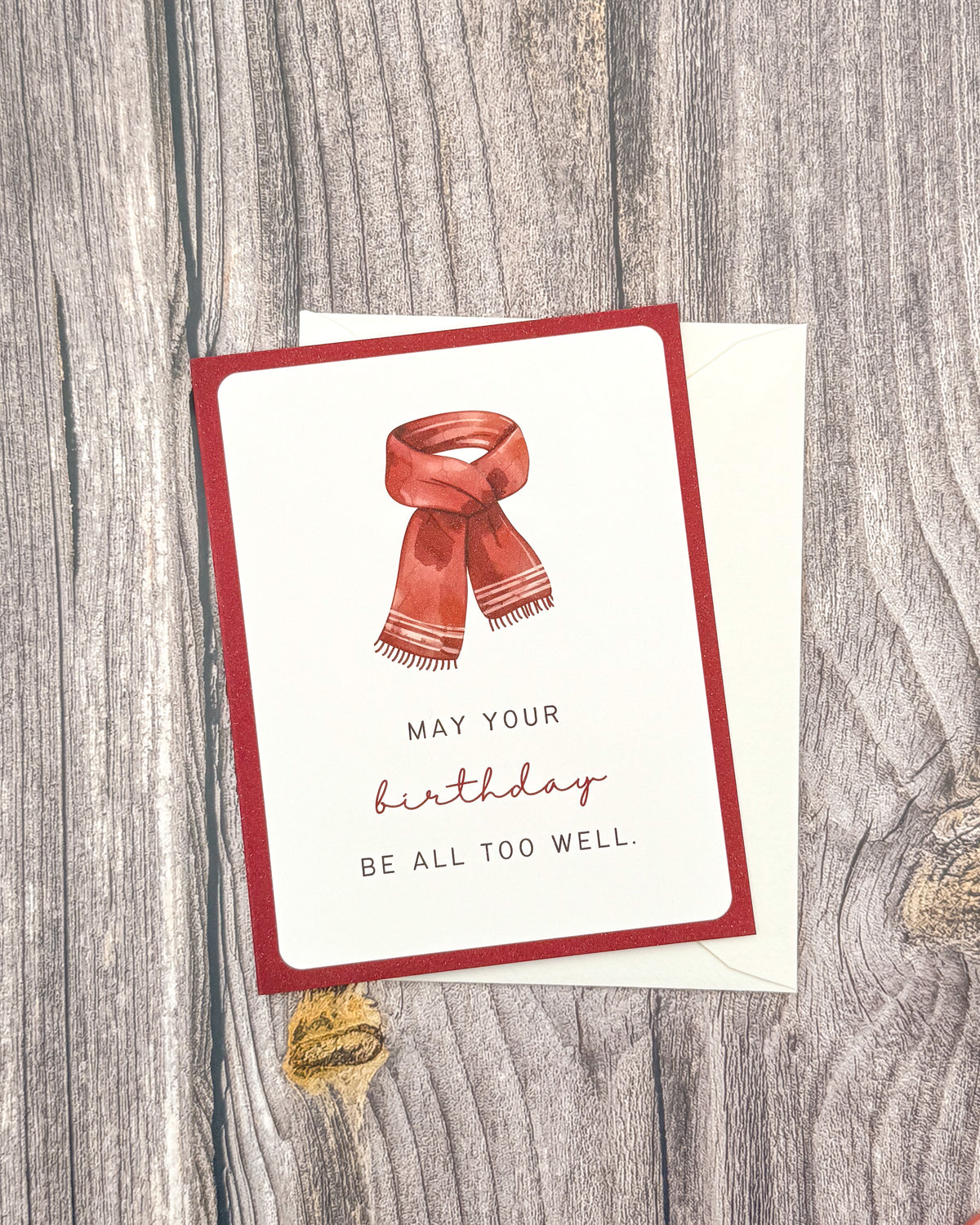 A square greeting card with a red border and a watercolor-style red scarf illustration, placed on top of a white envelope over a rustic gray wood surface. The card reads, 'MAY YOUR birthday BE ALL TOO WELL.