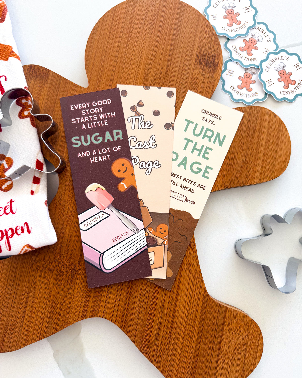 Three gingerbread-themed bookmarks with humorous quotes are arranged on a wooden gingerbread-shaped board, alongside cookie cutters and a white cloth with gingerbread text.