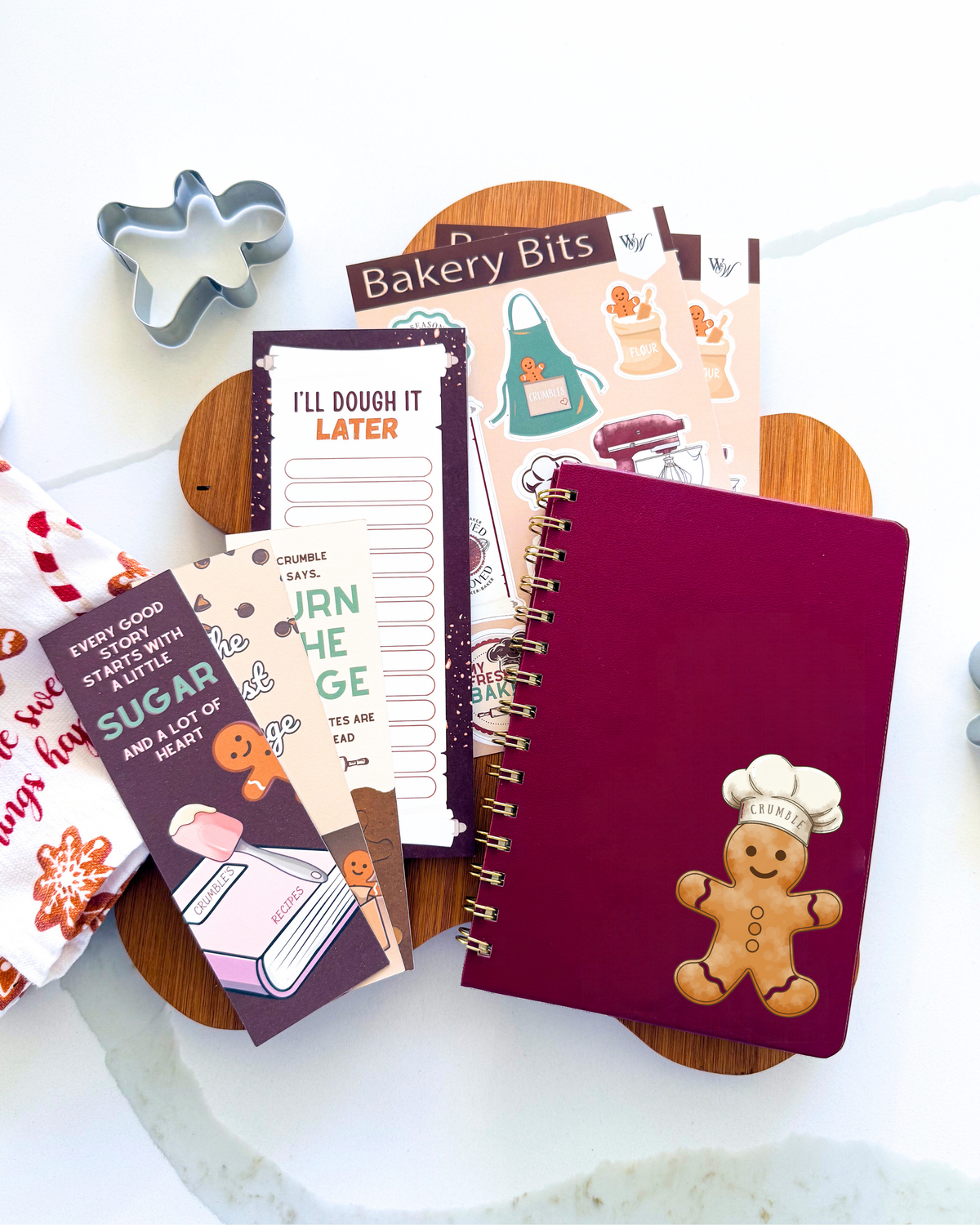 A styled flat-lay of gingerbread-themed stationery and baking tools on a white marble surface, including a deep burgundy spiral notebook with a gingerbread person chef, sticker sheets, cookie cutters, and baking notes.
