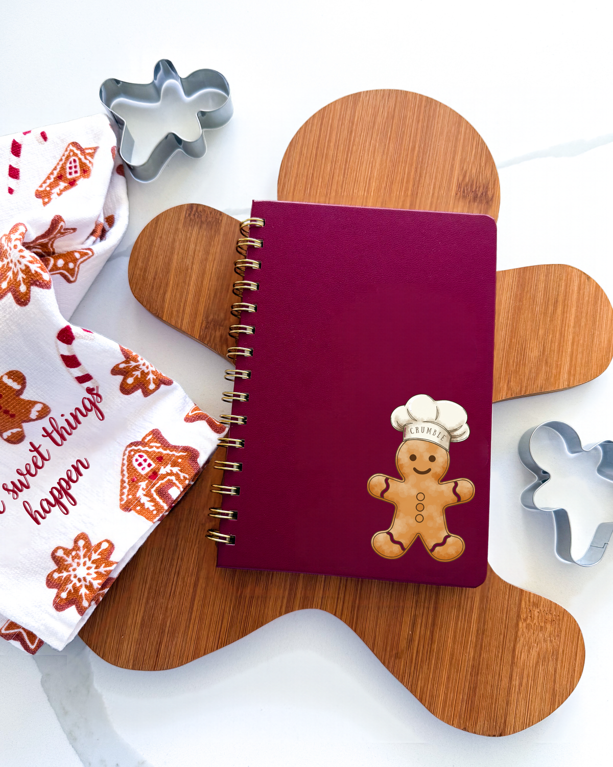 A burgundy spiral notebook with a gingerbread cookie decoration on the cover, styled on a wooden gingerbread man-shaped board with a white tea towel and metal cookie cutters.