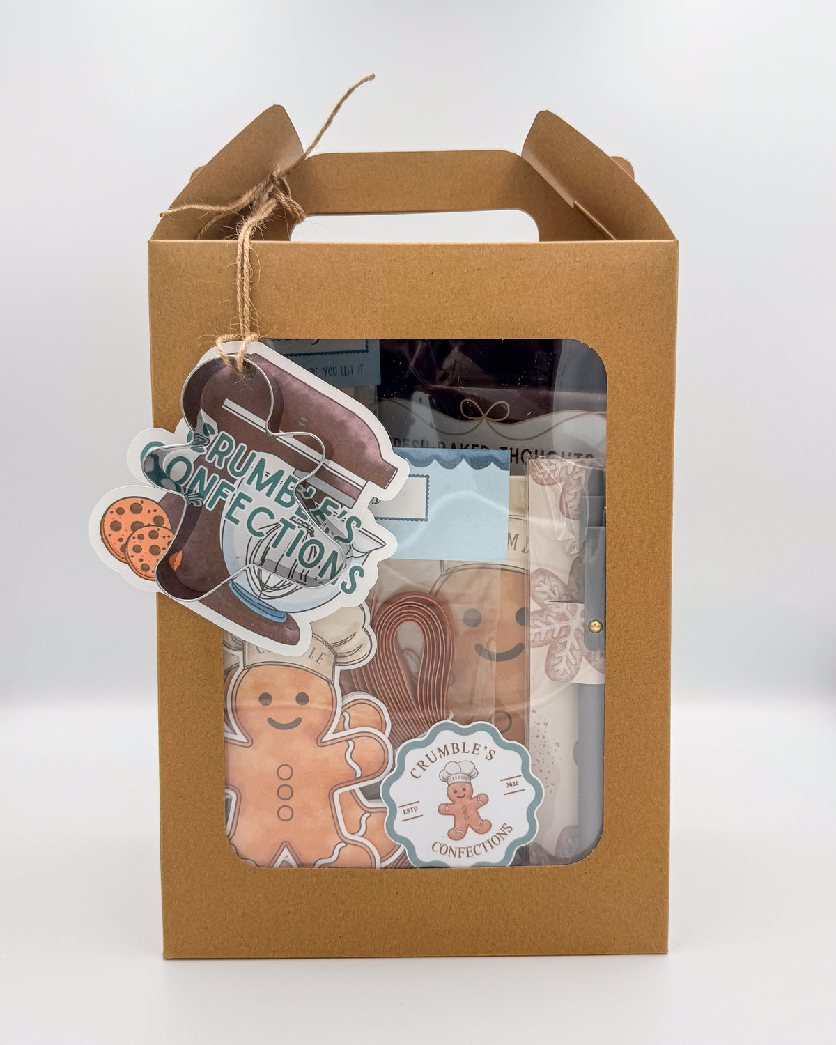 A brown kraft paper gift box with a clear window, containing assorted gingerbread-themed treats and packaged items. A hanging tag reads 'CRUMBLE'S CONFECTIONS' and shows gingerbread cookies and a gingerbread man illustration.