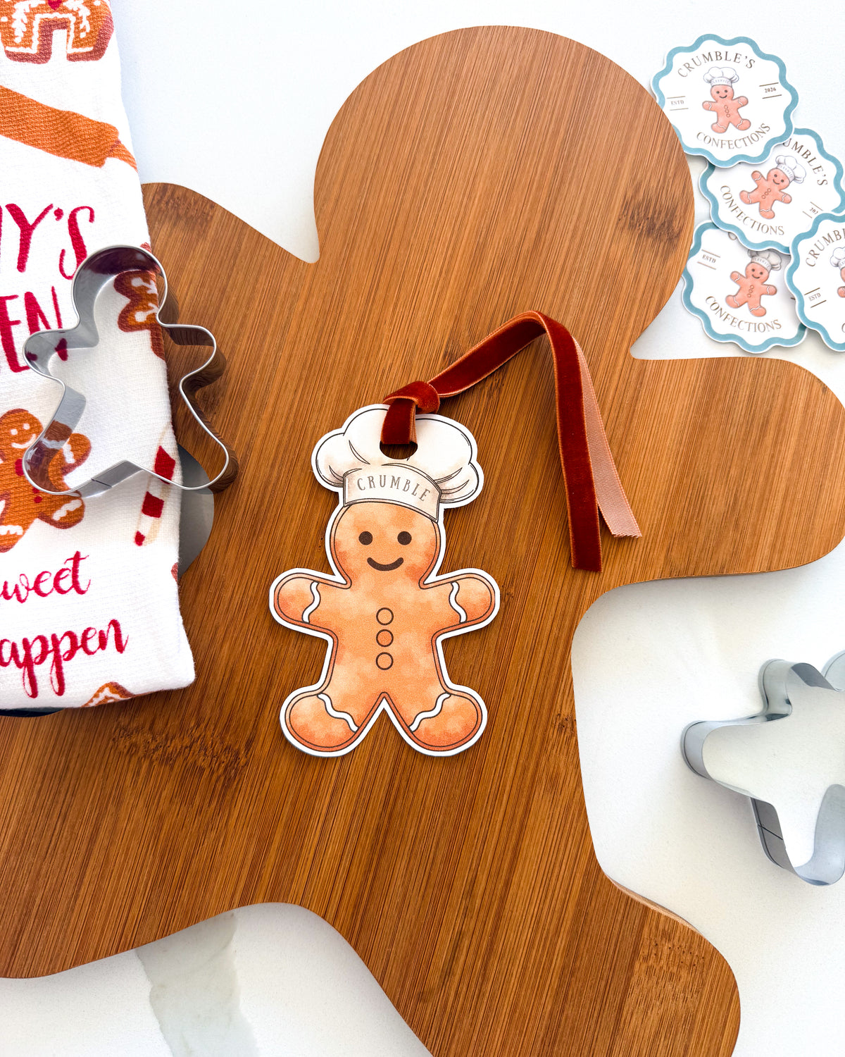 A gingerbread person ornament with a white chef hat and a red ribbon loop, displayed on a wooden cutting board with cookie cutters and a printed cloth nearby.