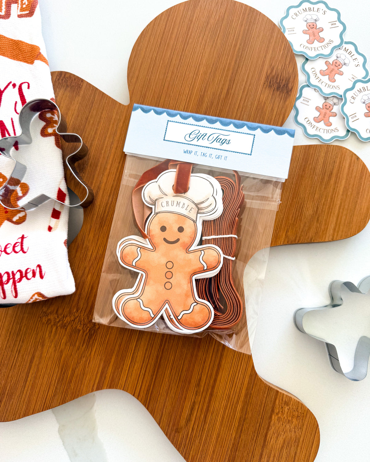 A packaged gingerbread cookie cutters set displayed on a wooden board, including a gingerbread person cutter, a white cloth with holiday text, and round decorative tags.