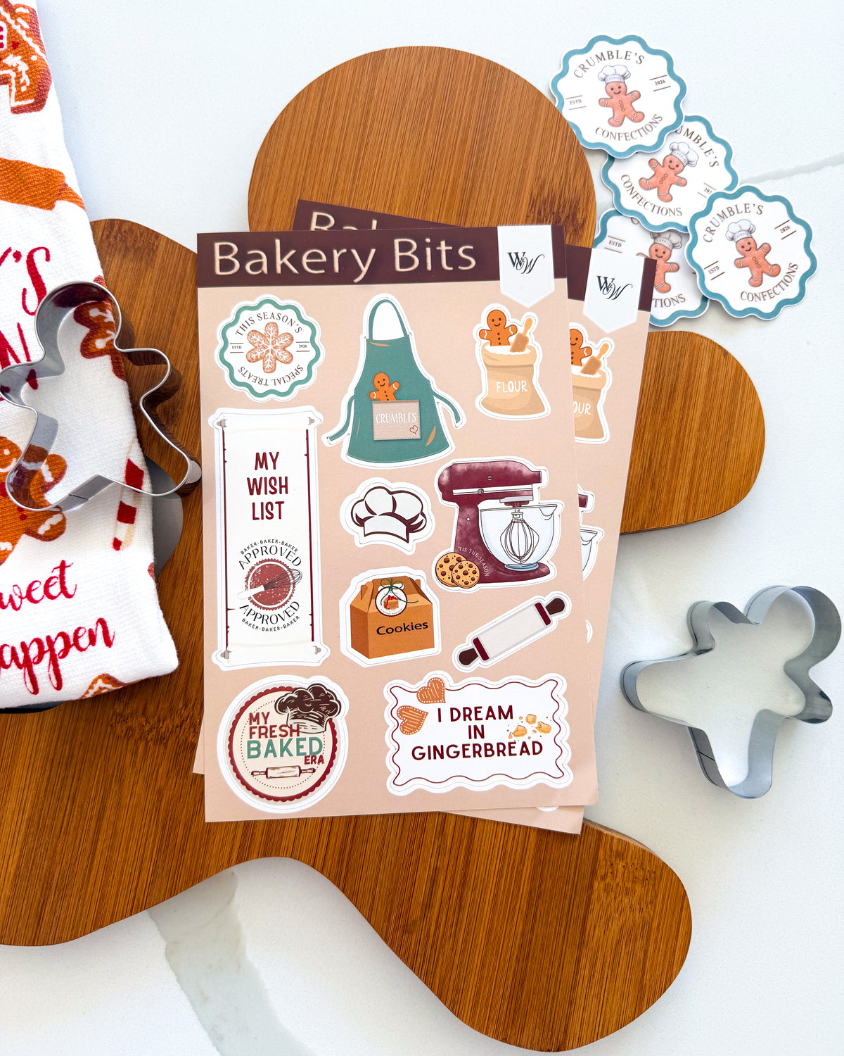 A sheet of decorative planner stickers labeled 'Bakery Bits' is displayed on a wooden gingerbread-shaped board, surrounded by baking-themed props including cookie cutters, a napkin, and matching sticker designs.