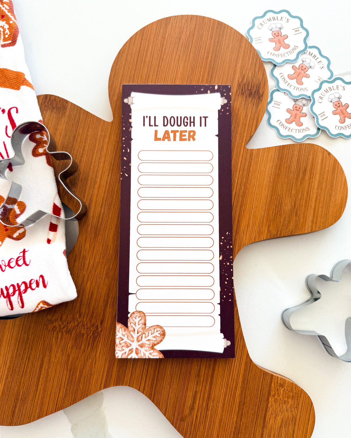 A gingerbread-themed notepad with the text 'I'LL DOUGH IT LATER' lies on a wooden gingerbread-shaped board, surrounded by cookie cutters and decorative gingerbread cookies on a white surface.