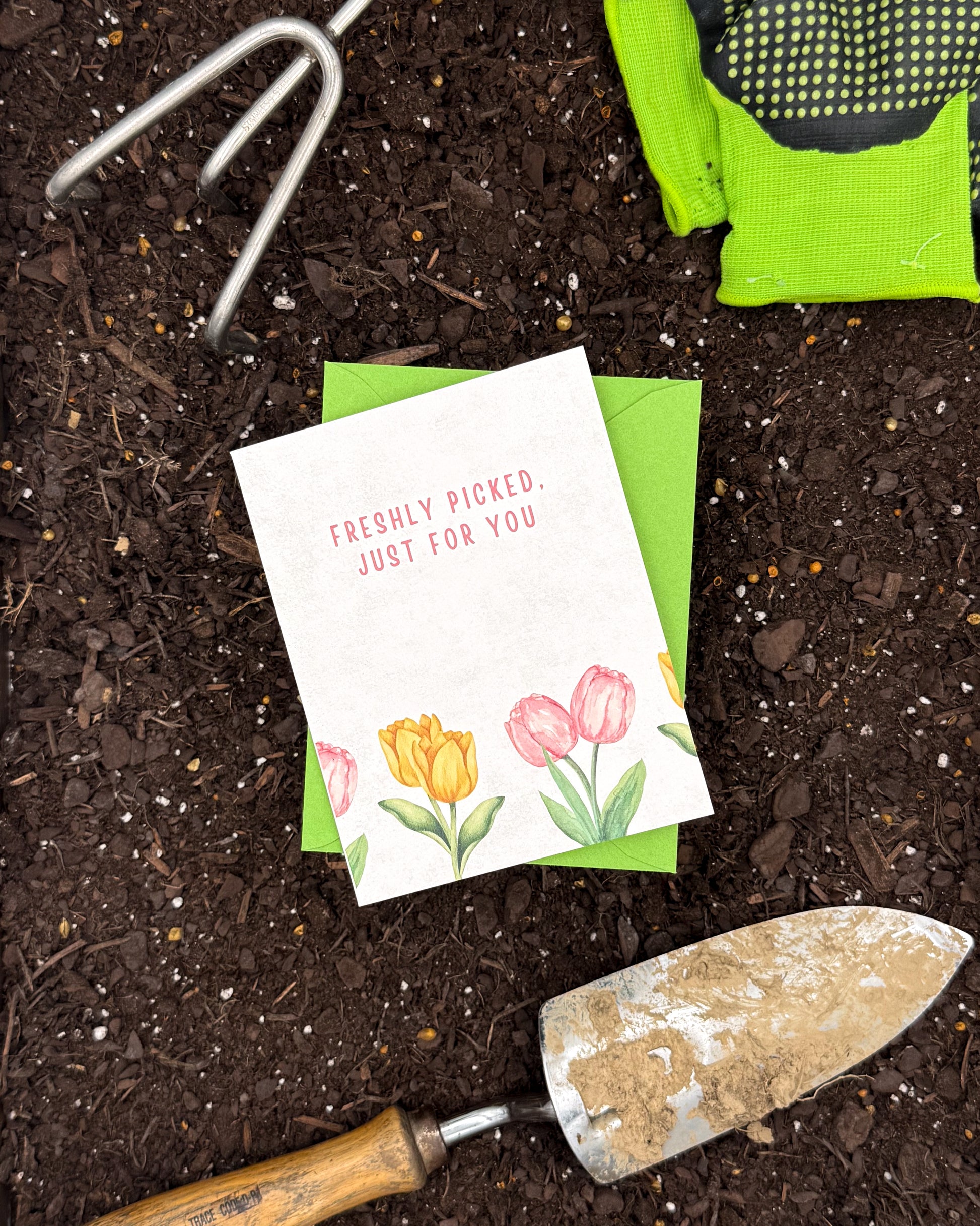 Gardening tools and a card with flowers on a bed of soil