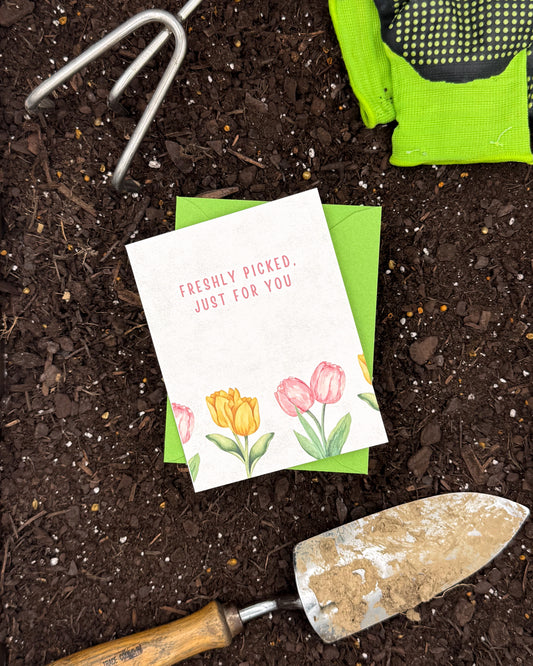 Gardening tools and a card with flowers on a bed of soil