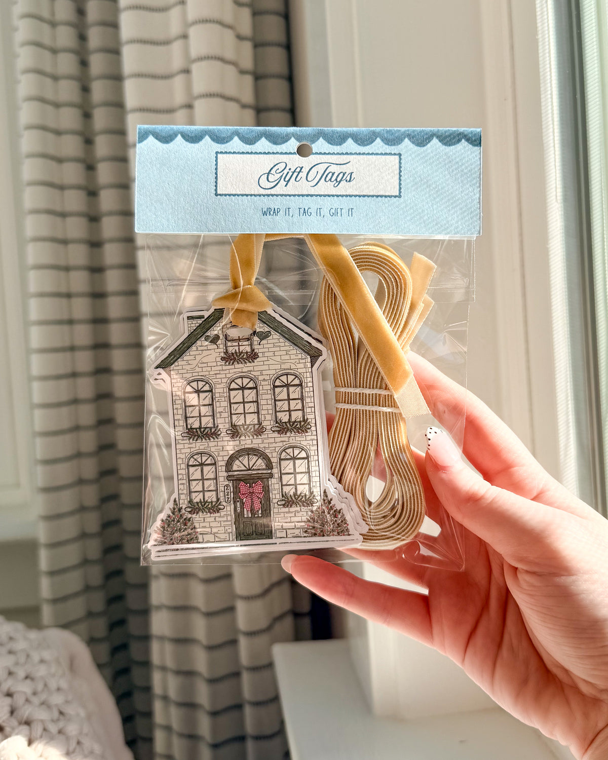 A hand holds a clear plastic bag of gift tags. The package contains tan ribbon loops and a decorative house-shaped tag with a black-and-white illustration and a small pink bow.