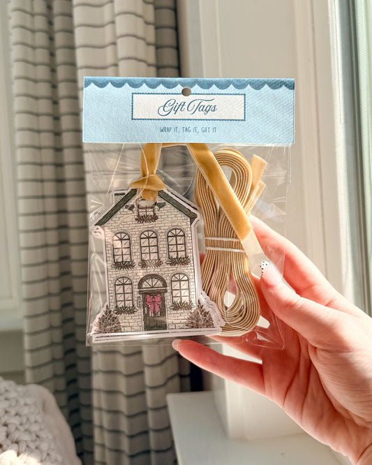 A hand holds a clear plastic bag of gift tags. The package contains tan ribbon loops and a decorative house-shaped tag with a black-and-white illustration and a small pink bow.