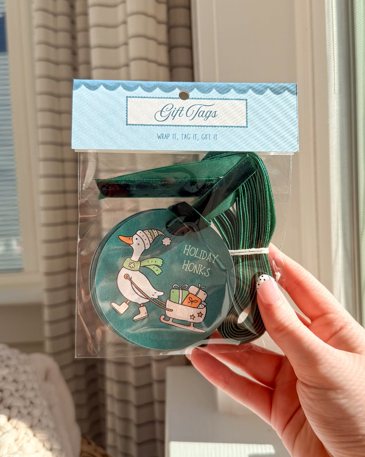 A hand holding a packaged set of holiday-themed gift tags. The set includes a round teal tag with a cartoon goose and the words 'HOLIDAYS HONORS,' plus several dark green rectangular tags behind it.