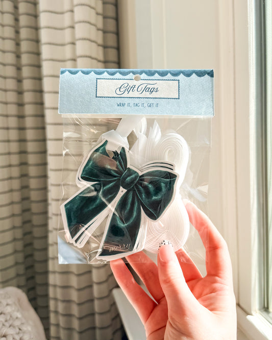 A hand holds a clear plastic package of white gift tags with a large dark green velvet bow on the front. The package has a light blue header card that reads 'Gift Tags'.