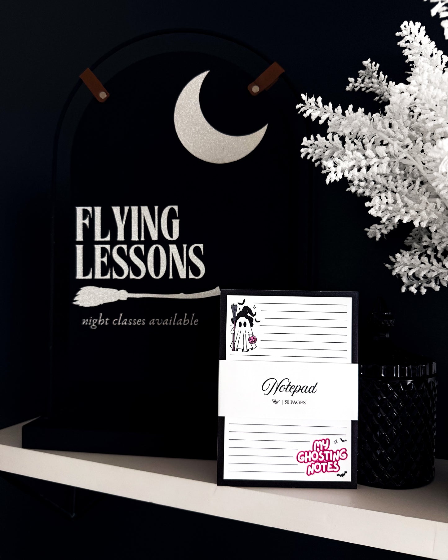 A black Halloween-themed wall sign reading 'FLYING LESSONS' with a broom and moon graphic, displayed on a shelf next to a matching notepad and white decorative foliage.