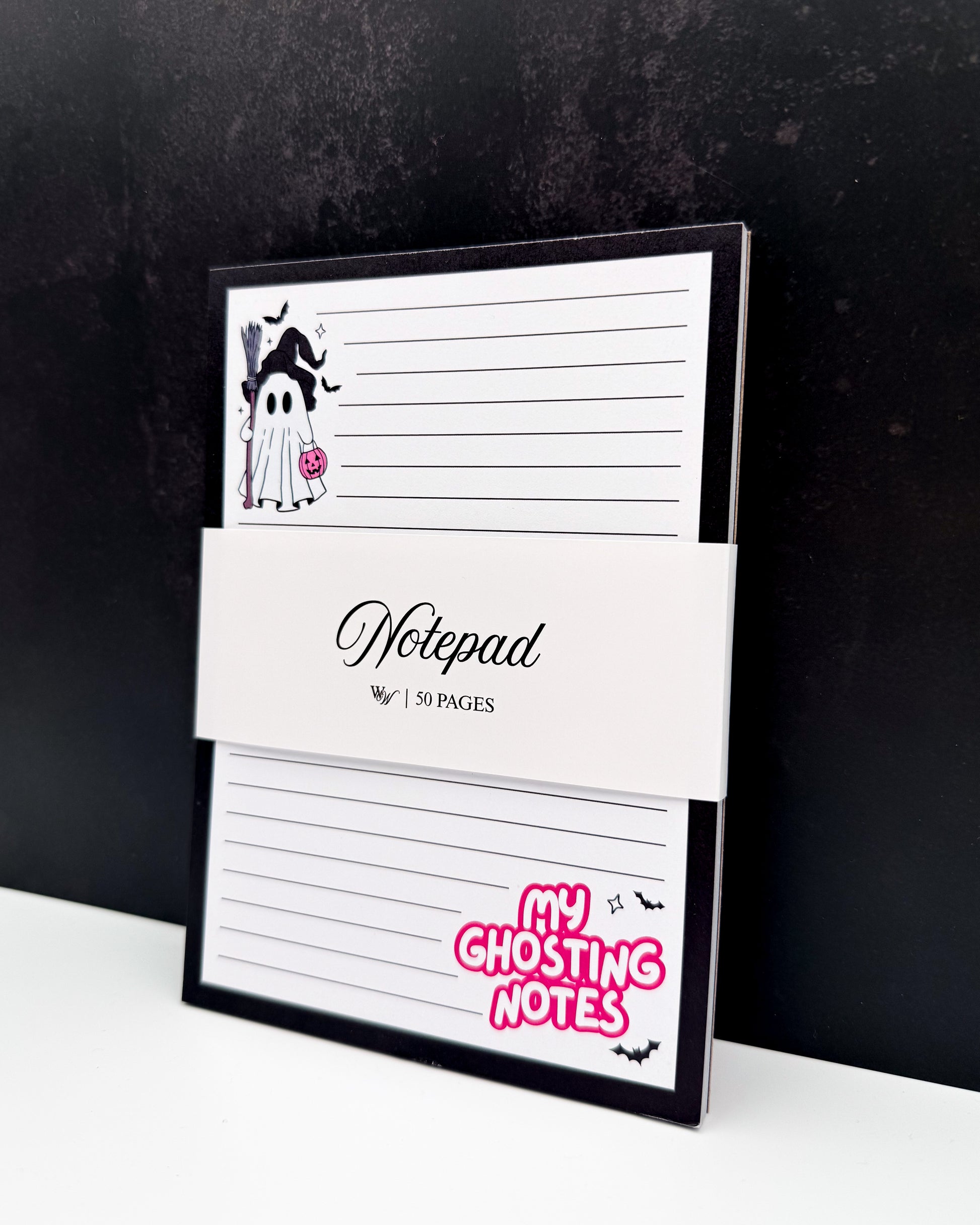 A Halloween-themed lined notepad with a black border, a white paper band reading 'Notepad' and '50 PAGES', and a pink 'MY GHOSTING NOTES' sticker on the bottom right. The notepad is photographed standing on a white surface against a dark textured background.