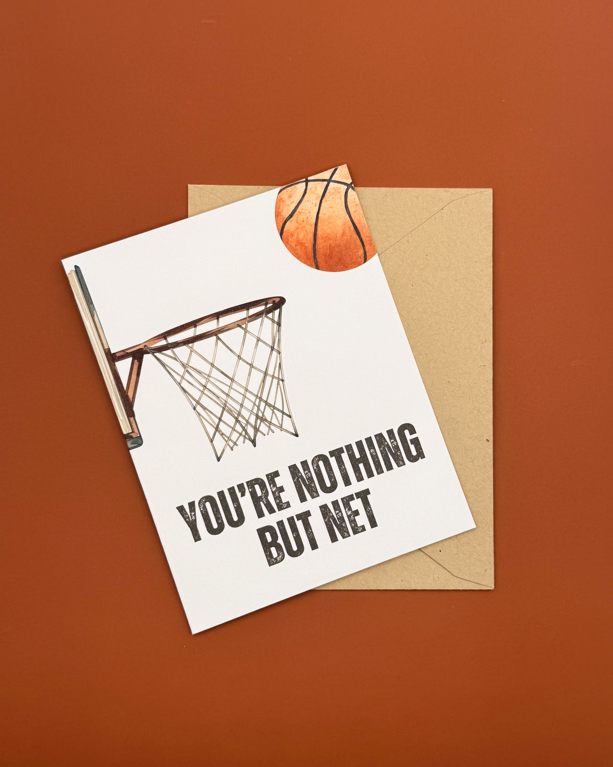 A white greeting card with a basketball hoop illustration and the text 'YOU'RE NOTHING BUT NET' lies on top of a kraft paper envelope against a solid terracotta background.
