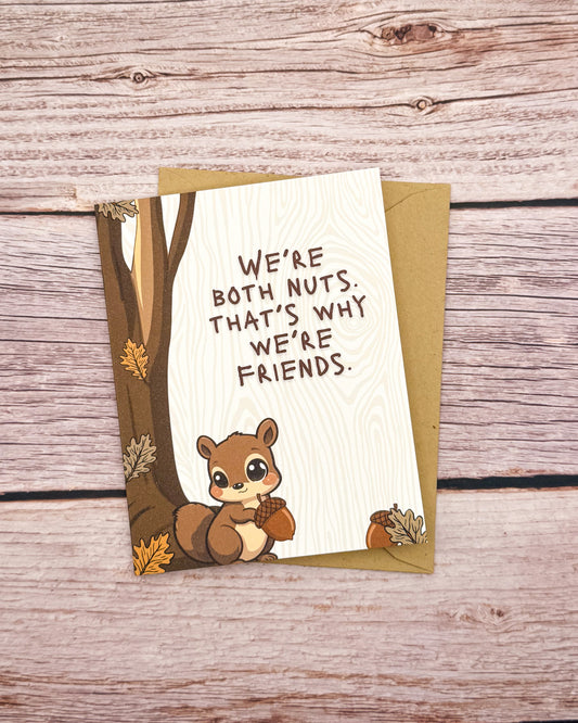 A cute autumn-themed greeting card featuring a cartoon chipmunk holding an acorn next to a tree trunk, with the text 'WE'RE BOTH NUTS. THAT'S WHY WE'RE FRIENDS.