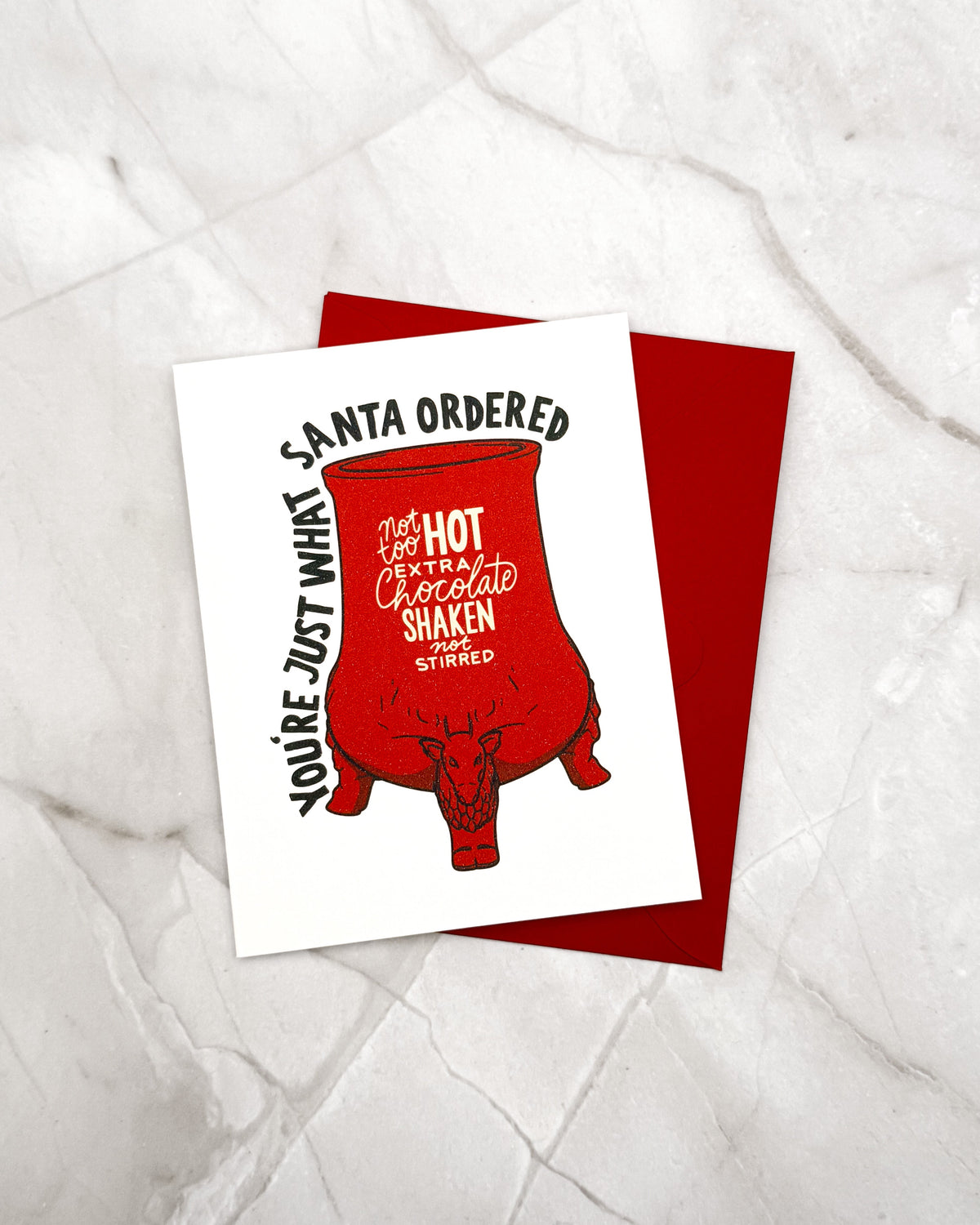 A white greeting card with a red mug illustration and black text reading 'SANTA ORDERED YOU'RE JUST WHAT YOU'RE JUST WHAT' lies on top of a red envelope, placed on a light marble surface.