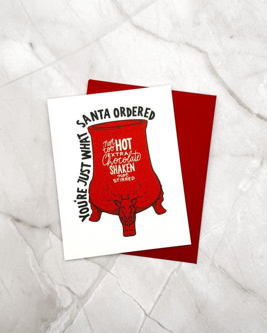 A white greeting card with a red mug illustration and black text reading 'SANTA ORDERED YOU'RE JUST WHAT YOU'RE JUST WHAT' lies on top of a red envelope, placed on a light marble surface.