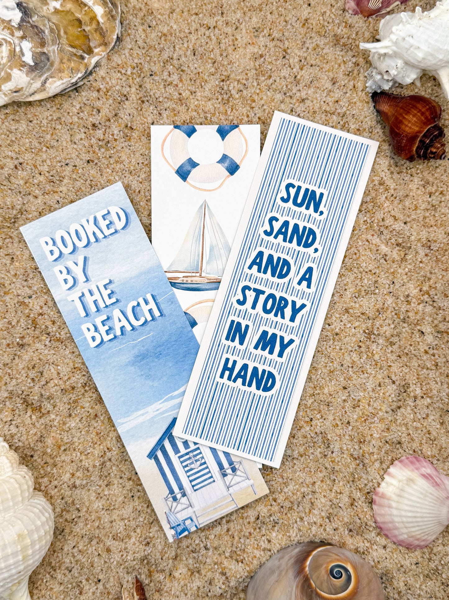 Three bookmarks with beach-themed designs on a sandy background with shells.