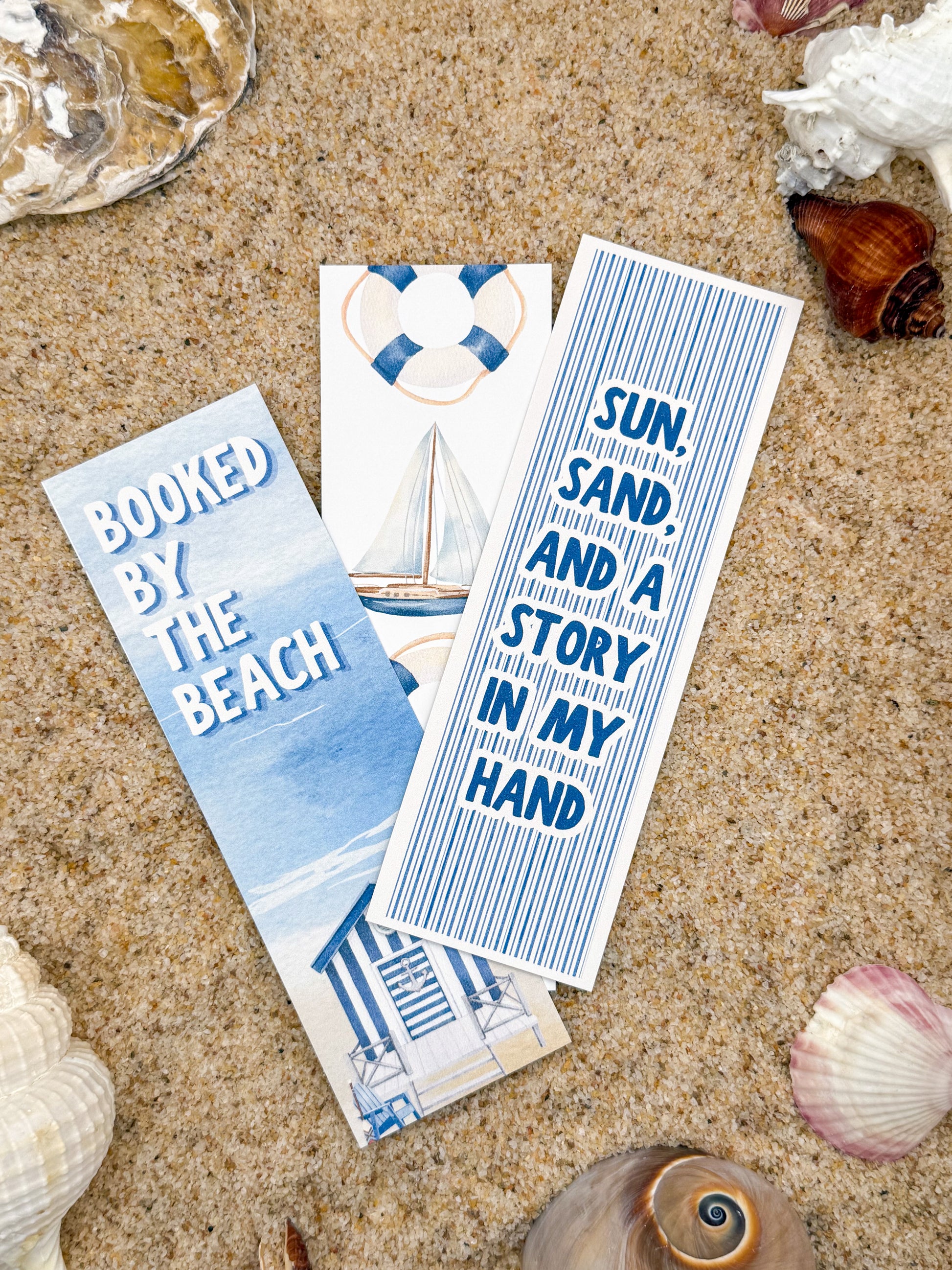 Three bookmarks with beach-themed designs on a sandy background with shells.