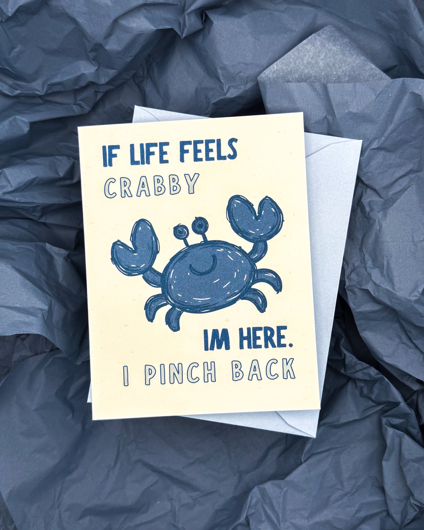 Card with a blue crab illustration and humorous text on a crumpled blue surface