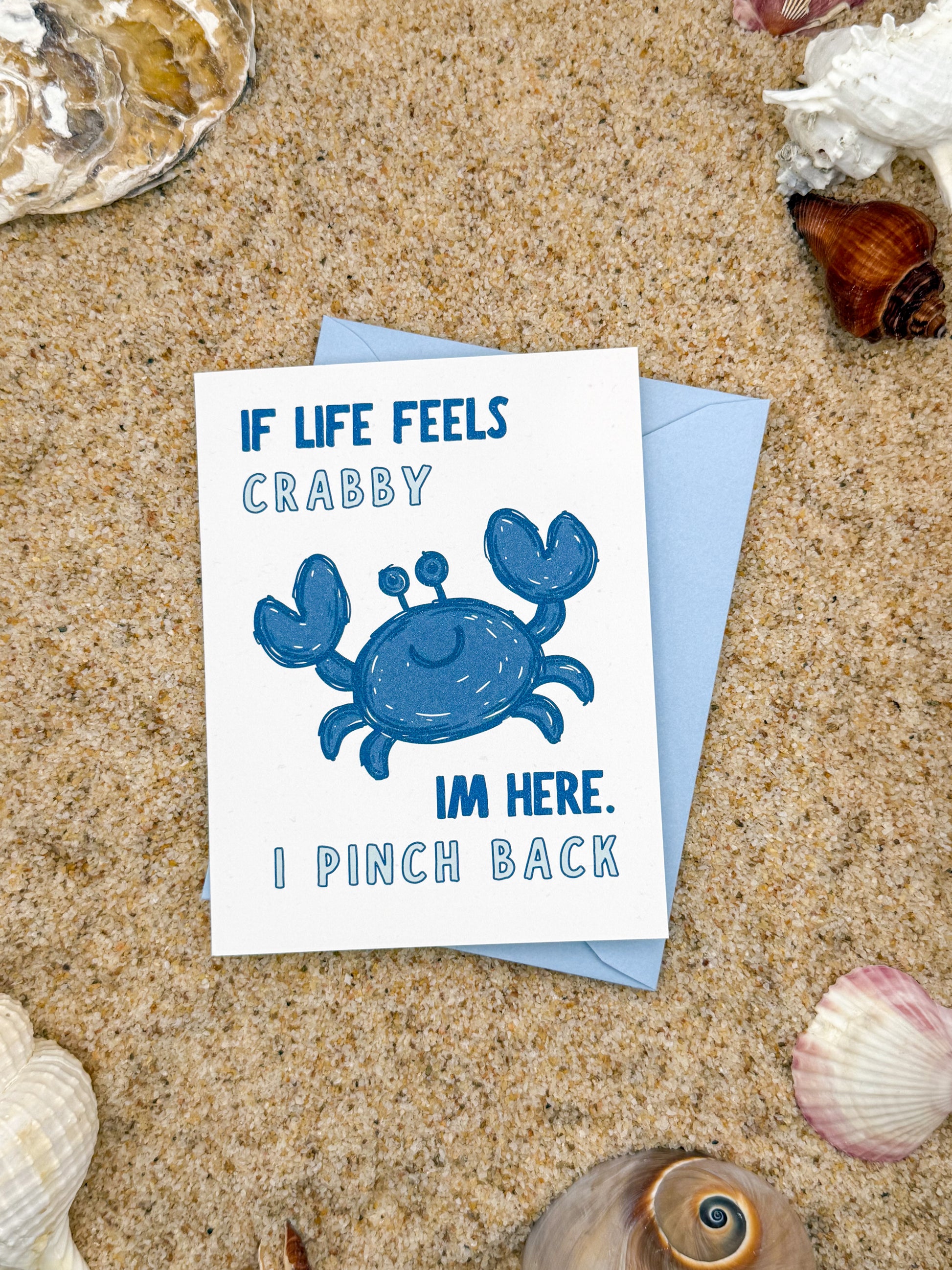 Card with a blue crab illustration and humorous text on sand with shells