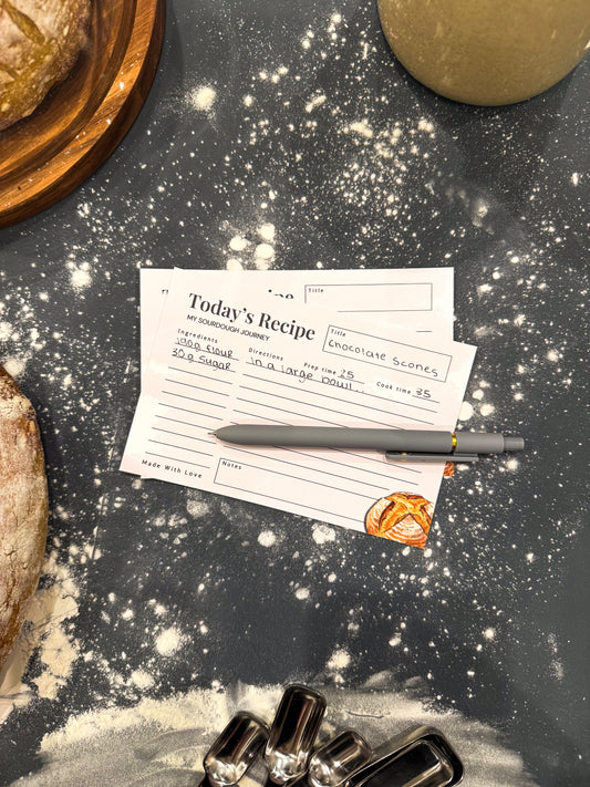 Recipe card with pen on a flour-dusted surface with bread and silver tongs.