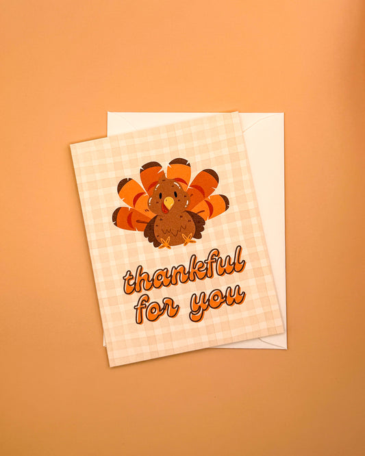 A Thanksgiving greeting card with a cartoon turkey and the words 'thankful for you' on a peach gingham background, shown with a white envelope on a peach background.