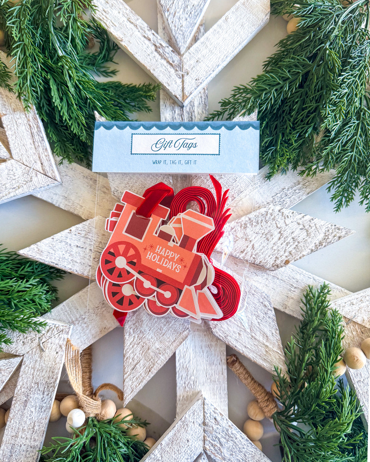 A festive Christmas ornament shaped like a red train with 'Happy Holidays' written on it, displayed on a rustic white star wreath with green pine branches and a small gift tag reading 'Gift Tags'.