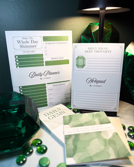 Green and white stationery items including a daily planner, notepad, and gem-themed books on a dark surface.