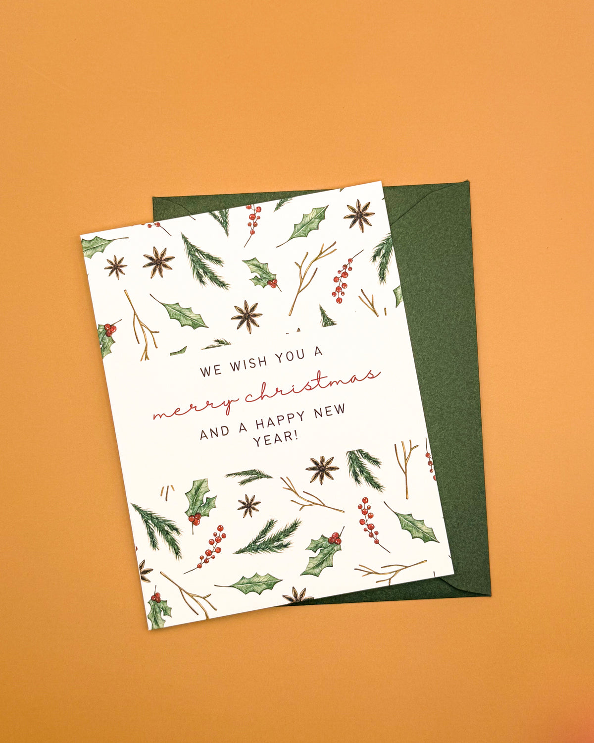 A holiday greeting card with a white background and botanical illustrations of holly leaves, berries, and starbursts, placed on top of a dark green envelope against a solid orange background.