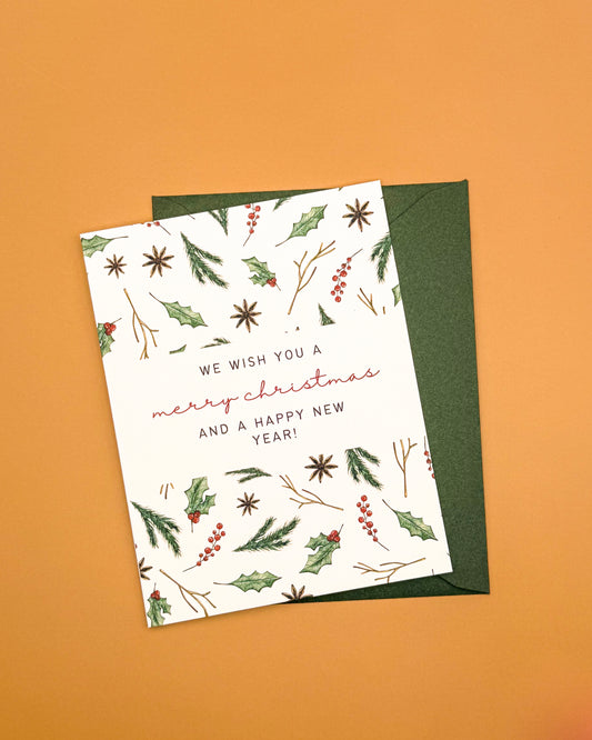 A holiday greeting card with a white background and botanical illustrations of holly leaves, berries, and starbursts, placed on top of a dark green envelope against a solid orange background.