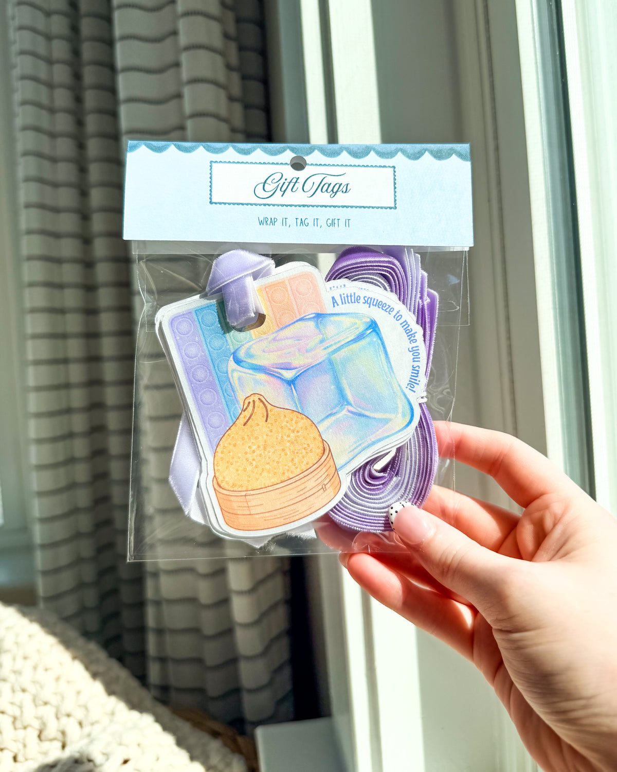 A hand holding a clear plastic bag of pastel gift tags with a light blue header card. The tags feature illustrated food and drink designs in lavender, blue, purple, and peach tones, photographed in front of a window with curtains.