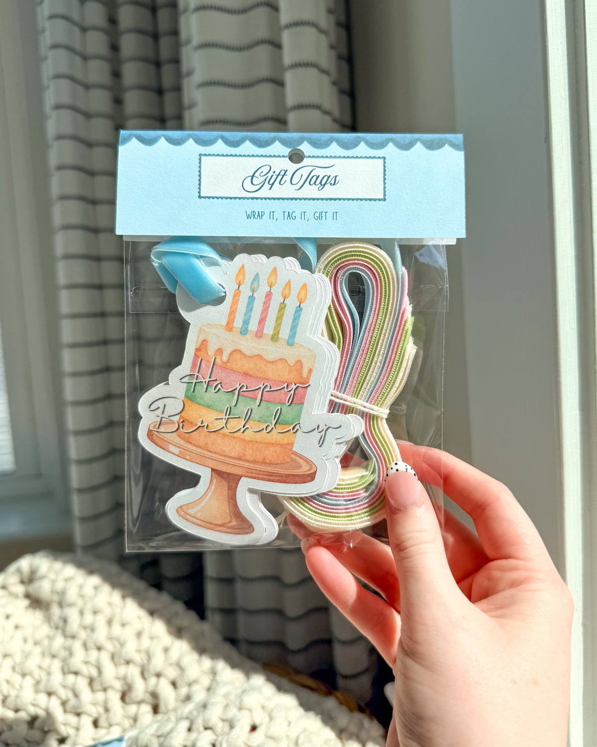 A hand holds a packaged set of gift tags featuring a birthday cake illustration and a rainbow-colored loop string, with a light blue header card that reads 'Gift Tags'.
