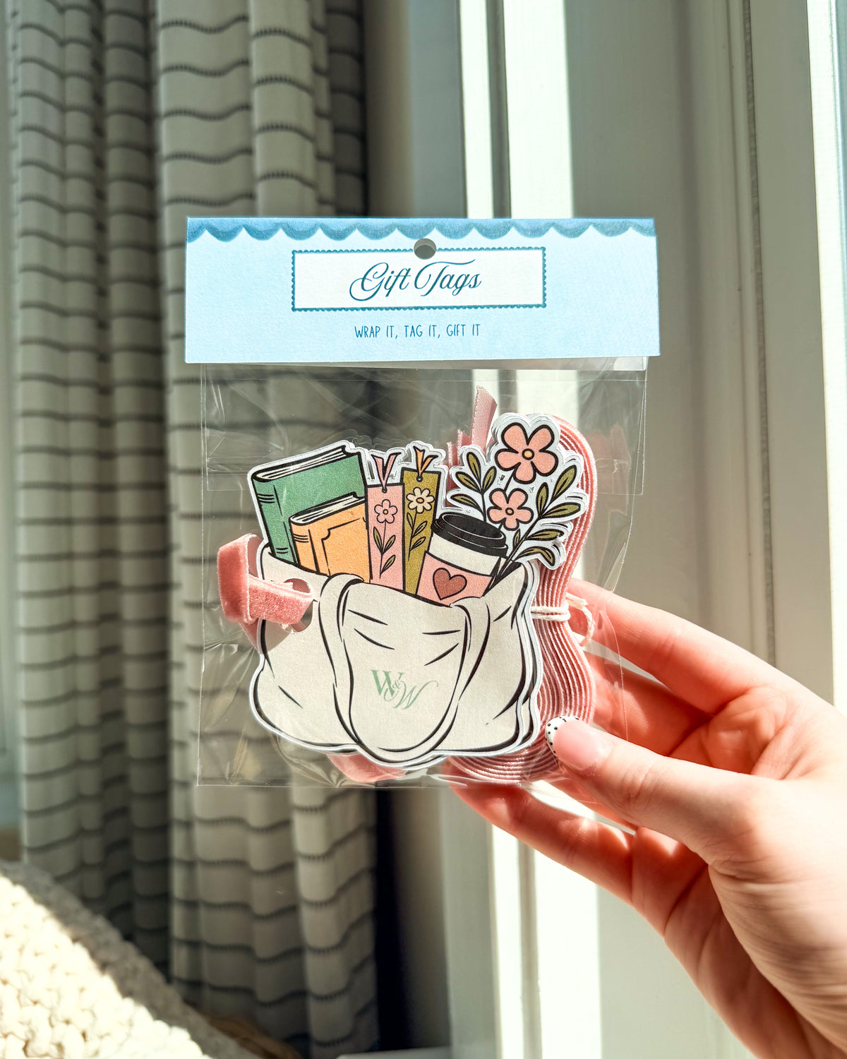 A hand holds a clear plastic bag containing a set of decorative gift tags. The tags feature a white fabric pouch illustration with the word 'Wish' embroidered on it, surrounded by colorful books, flowers, and a coffee cup.