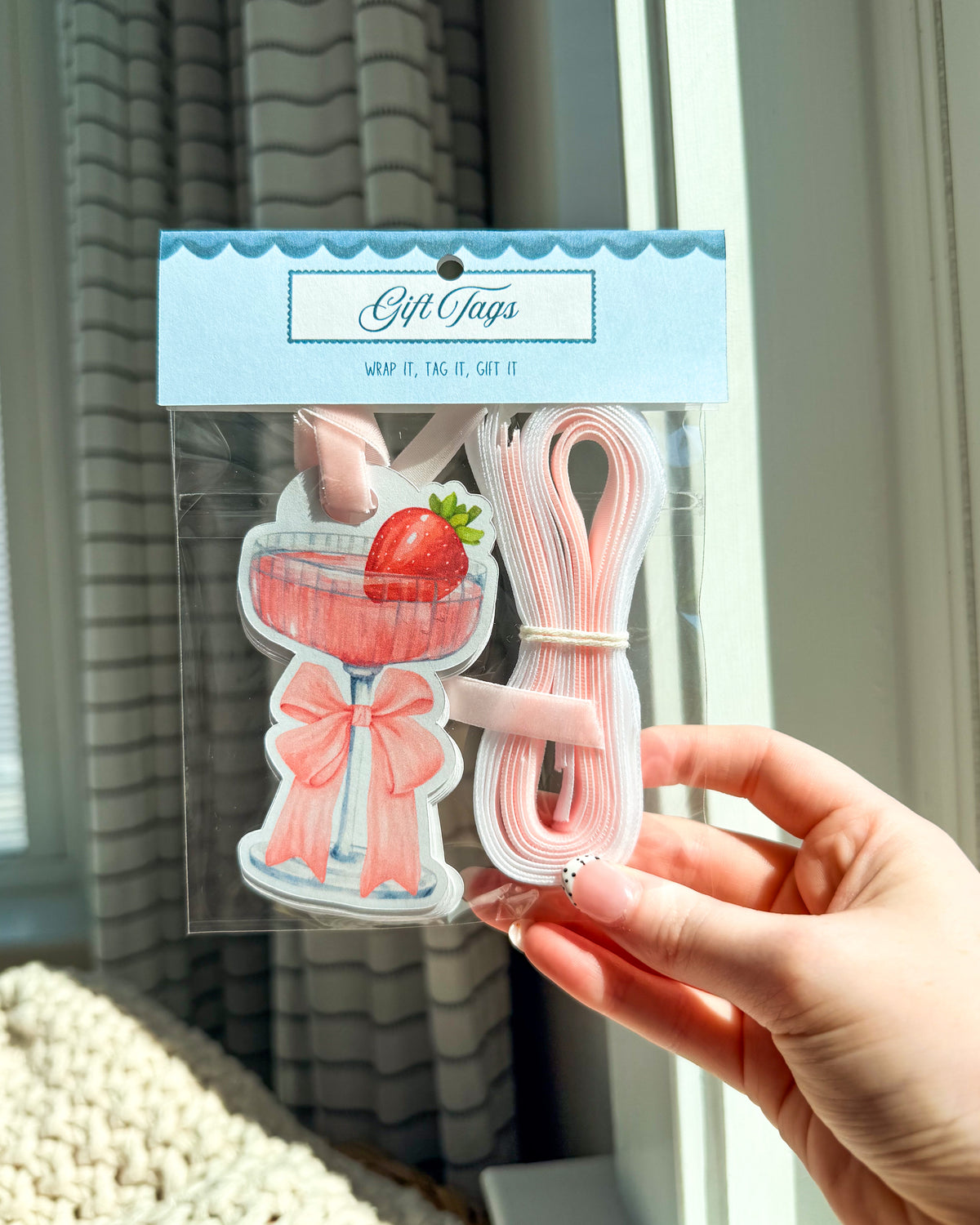 A hand holds a packaged set of gift tags shaped like a pink cocktail glass with a strawberry and a pink bow, alongside a roll of pink elastic bands, against a window background.
