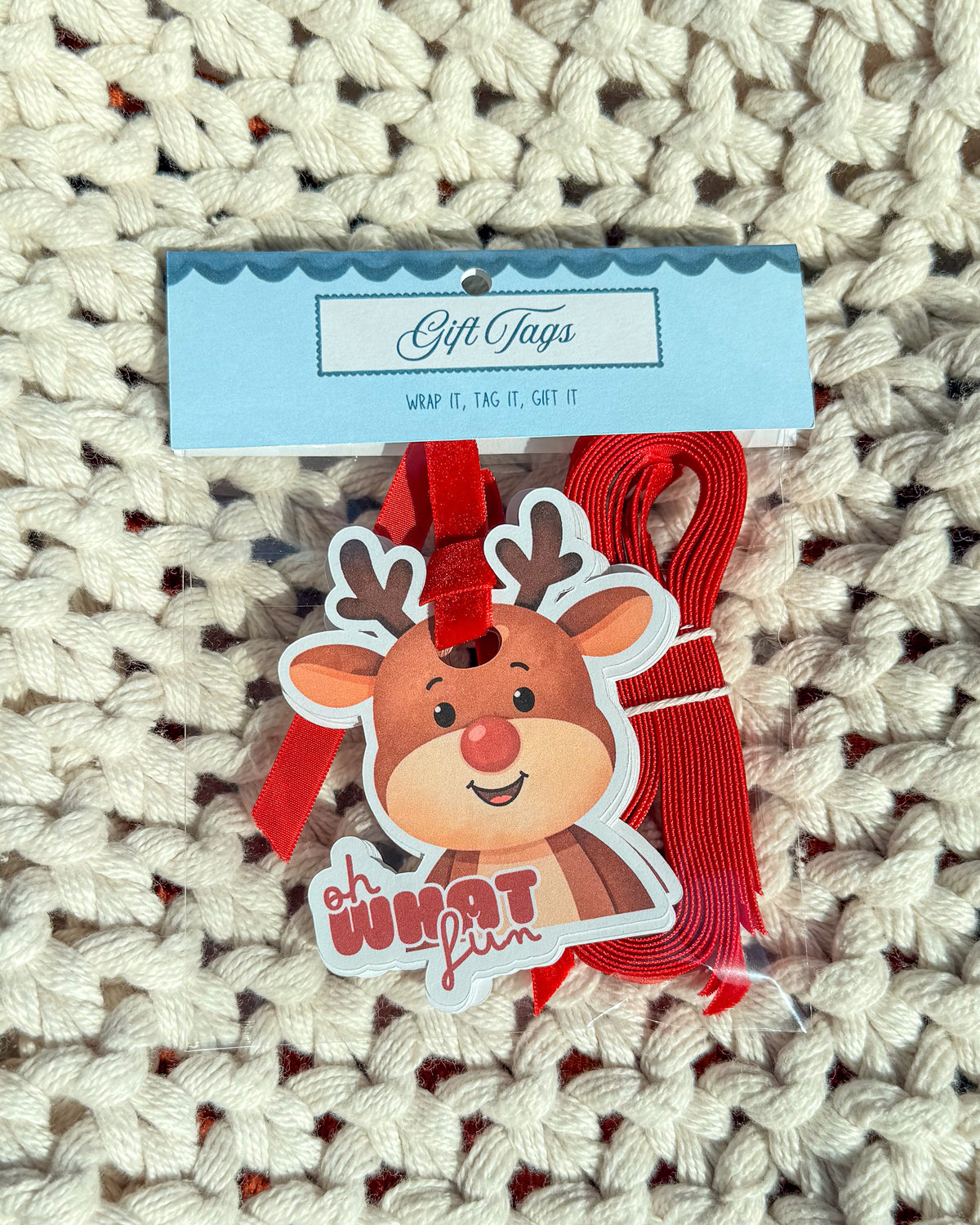 A Christmas-themed gift tag set featuring a die-cut cartoon reindeer sticker with the phrase 'Oh What Fun' and a red ribbon loop, attached to a light blue card labeled 'Gift Tags' and placed on a cream knit blanket.
