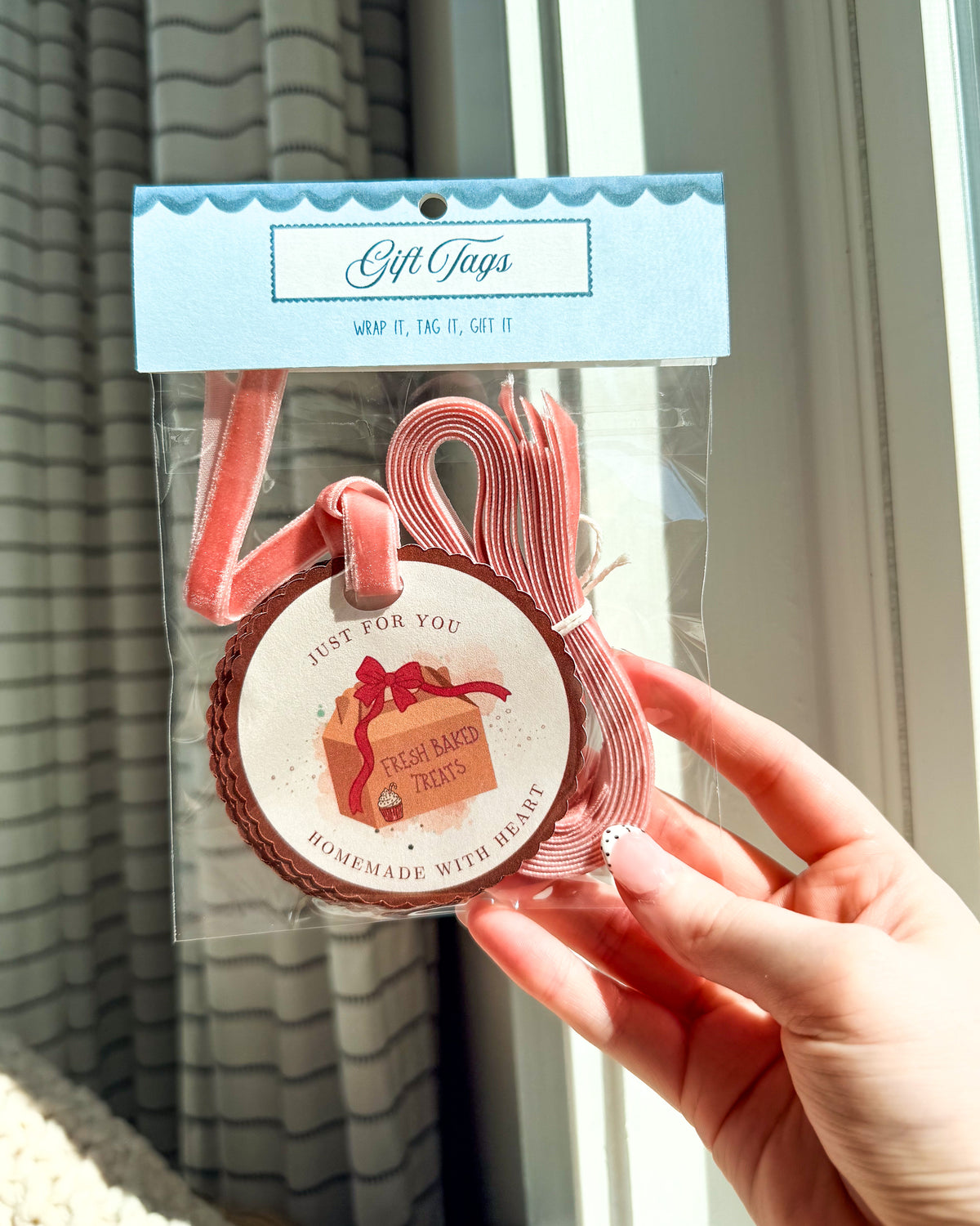 A hand holding a packaged set of pink gift tags. The tags are attached to a clear plastic bag with a light blue header card labeled 'Gift Tags' and a round paper tag that reads 'Just for you Fresh Baked Treats Homemade with Love.